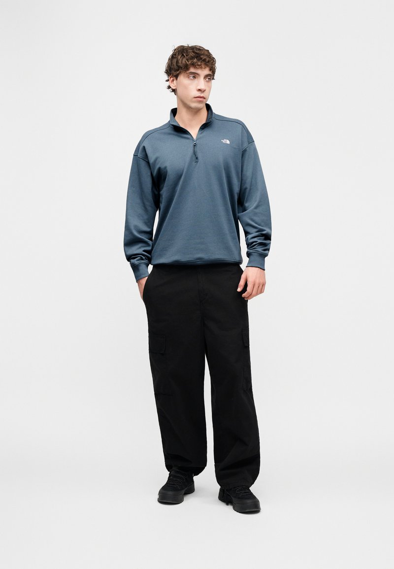 Blue long-sleeve pullover with a zip neck, made of soft fabric, paired with black cargo pants featuring side pockets, worn with black shoes.