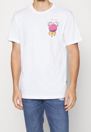 T-shirt blanc à manches courtes avec un petit graphique coloré sur la poitrine représentant des mains griffues tenant un cercle rose avec le texte "Nike", porté avec un jean bleu.