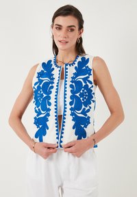 White embroidered vest featuring blue floral patterns, a front zipper, and decorative scalloped edges. Worn over a white top.