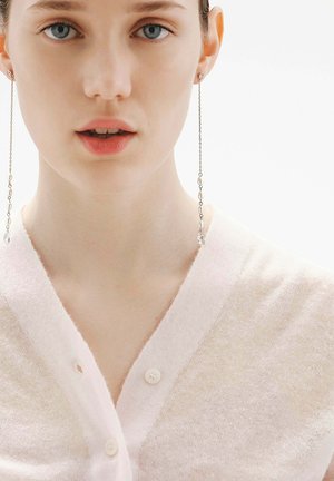 Primer plano de una mujer que lleva pendientes largos y delicados de cadena con pequeñas cuentas, vestida con un cárdigan abotonado de color claro y suave.