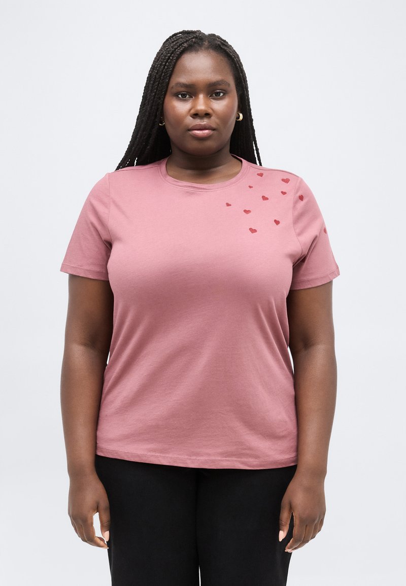 Donna con lunghe trecce che indossa una maglietta rosa polvere con piccoli disegni di cuori rossi sulla spalla, in piedi contro uno sfondo chiaro e uniforme.