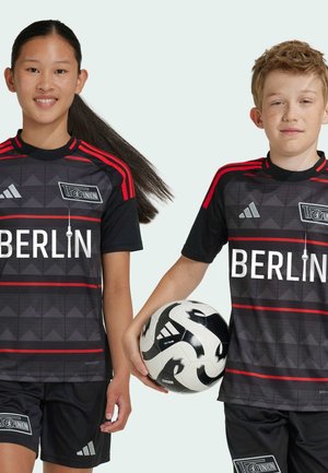 Schwarze Fußballtrikots mit roten Akzenten und dreieckigen Mustern, mit "BERLIN" gut sichtbar. Ein Schwarz-Weiß-Fußball wird gehalten.