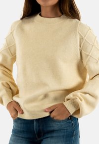 Maglione a maglia color crema con scollatura rotonda, maniche testurizzate caratterizzate da un motivo a diamante e dettagli in perla. Abbinato a jeans blu.