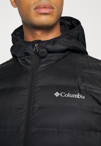 Giacca nera isolata con cappuccio, caratterizzata da una cerniera completa, design trapuntato orizzontale e un logo Columbia argentato sul petto.