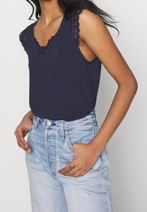 Kobieta w granatowej bluzce bez rękawów z koronkową lamówką i jasnoniebieskich wysokich dżinsach, jedna ręka w kieszeni, z długimi czarnymi włosami unoszącymi się na wietrze.