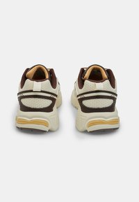 Vista posteriore di scarpe da ginnastica beige e marroni con pannelli in rete, suole in gomma testurizzata e accenti a contrasto. Presentano un colletto imbottito e cuciture.