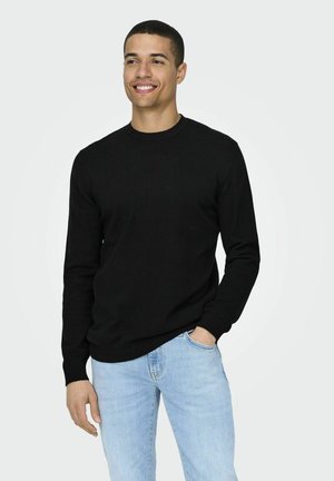 Un pullover noir à col rond en tissu doux, avec des manches longues et des poignets côtelés. Associé à un jean bleu clair, mettant en avant un look décontracté.