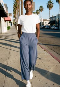 Tânăr mergând pe o trotuar însorită mărginită de palmieri, purtând tricou alb, pantaloni largi albaștri și adidași albi.