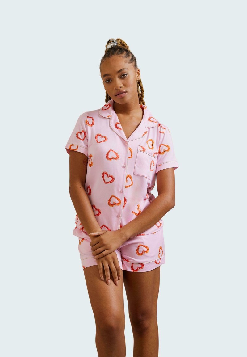 Pink pajama set with short sleeves and shorts, featuring a heart pattern in red and orange. Smooth fabric with a relaxed fit and pocket detail.