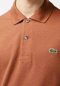 Brun polo skjorta i texturerat tyg med en tvåknapps knäppning. Har en grön broderad krokodil-logotyp på vänster bröst.