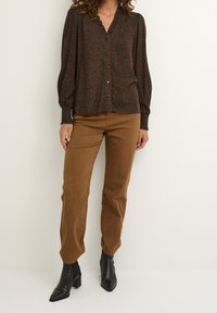 Woman wearing brown tapered pants, black heeled ankle boots, and a dark brown button-up blouse with a red floral pattern.