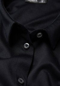 Schwarzes Hemd mit glatter Textur, Button-Down-Kragen und zwei grauen Kunststoffknöpfen auf der Vorderseite. Etikett am Kragen sichtbar.