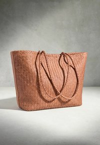 Grand sac fourre-tout en cuir tressé marron clair avec deux anses d'épaule tressées sur une surface claire et un fond gris.
