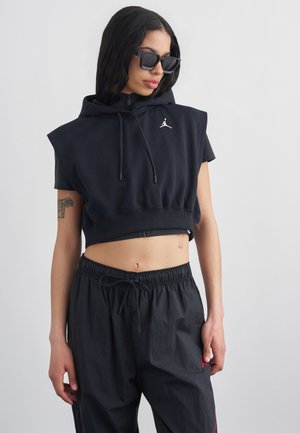 Femeie purtând hanorac negru scurt fără mâneci și pantaloni negri largi cu șnur, privind în jos, purtând ochelari de soare negri mari pe un fundal simplu.