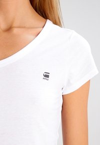Biała bawełniana koszulka z dekoltem w serek i krótkimi rękawami, z małym szarym logo "RAW" na lewej piersi. Gładka tekstura.