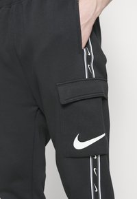 Mustat collegehousut, joissa on sivutasku, valkoinen Nike-logo ja valkoiset sivusuunnat, joissa on toistuvia Nike-logoja. Pehmeä, teksturoidusta kankaasta.