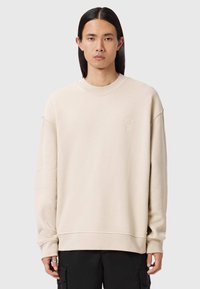 Giovane con lunghi capelli neri indossa una felpa beige oversize e pantaloni cargo neri, in piedi contro uno sfondo grigio chiaro semplice.