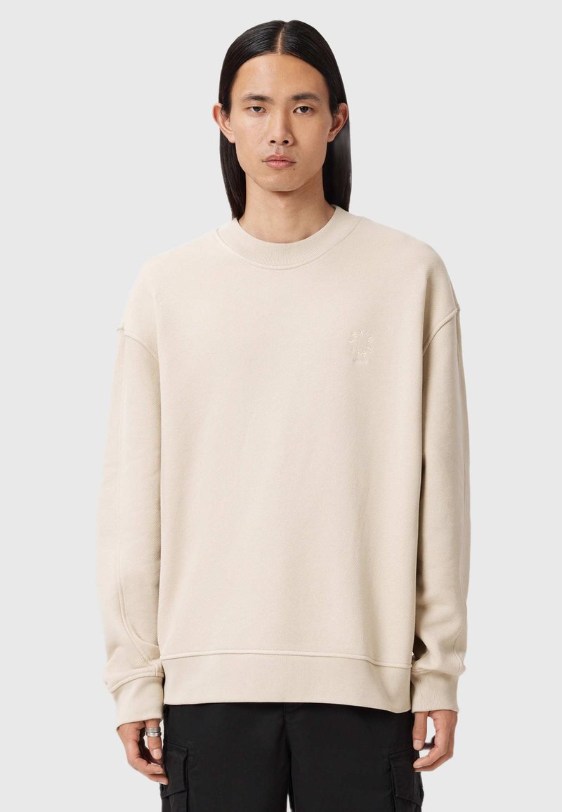 Giovane con lunghi capelli neri indossa una felpa beige oversize e pantaloni cargo neri, in piedi contro uno sfondo grigio chiaro semplice.