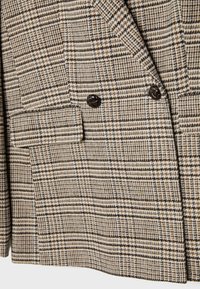 Veste en tissu à carreaux marron et beige avec des boutons noirs et une poche à rabat montrée en gros plan.