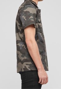Camicia a maniche corte in camo con tonalità di verde, marrone e nero smorzate; presenta un colletto con bottoni e una tasca sul petto; realizzata in tessuto misto cotone.
