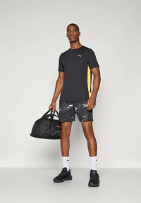 Must have mustekasärgi, millel on kollased külje paneelid, koos mustade ja hallide mustritega lühikeste pükstega. Must duffel-kott, mustad kingad ja valged sokid.