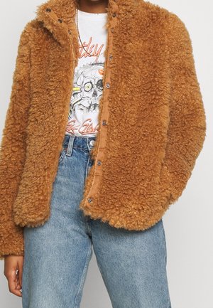 Person wearing brown curly faux fur jacket over white graphic skull and text T-shirt, paired with high-waisted faded blue jeans.