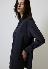 Camicia a maniche lunghe color navy realizzata in tessuto liscio, con chiusura frontale a bottoni e polsini annodati, dalla silhouette morbida e una leggera lucentezza.