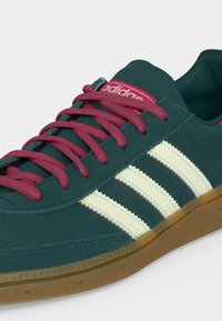 Πράσινο παπούτσι Adidas με λευκές ρίγες, κόκκινα κορδόνια, κόκκινο λογότυπο στη γλώσσα και διαφανές καφέ λαστιχένιο πέλμα, εμφανίζεται από πλάγια γωνία.
