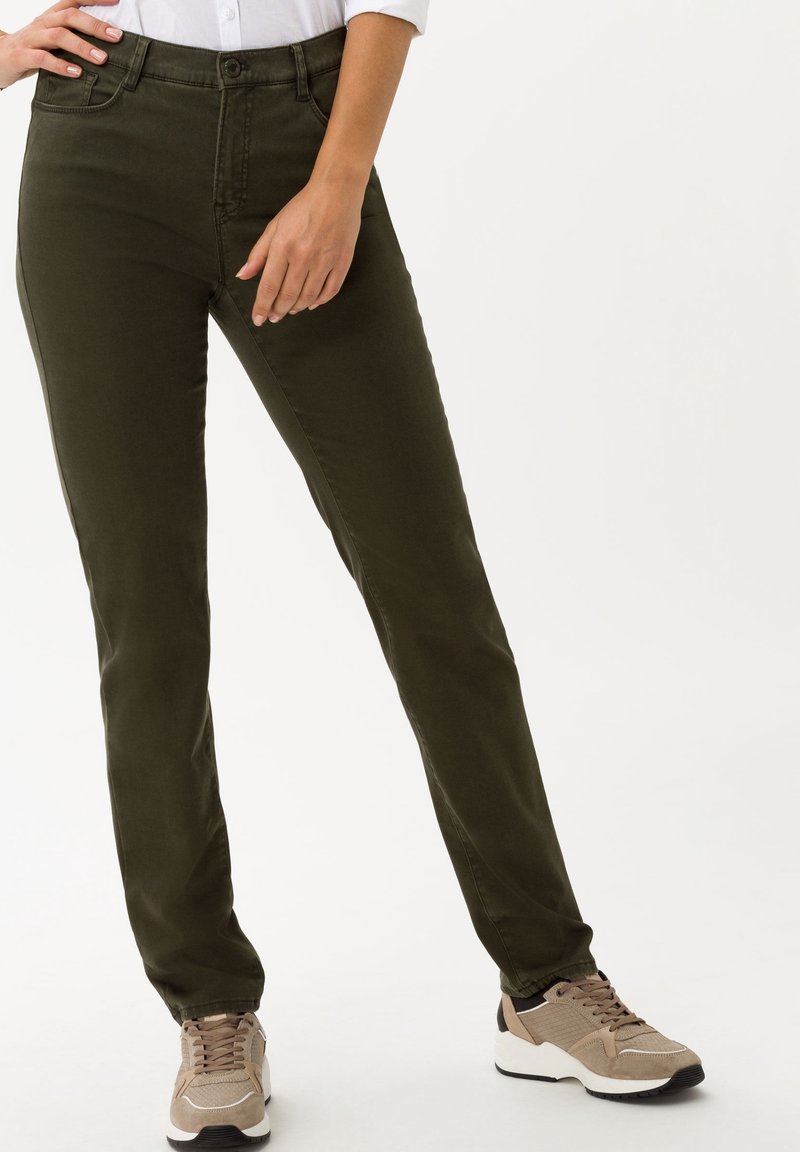 Pantalon slim-fit kaki foncé porté avec des baskets beige et marron sur une personne debout devant un fond clair uni.