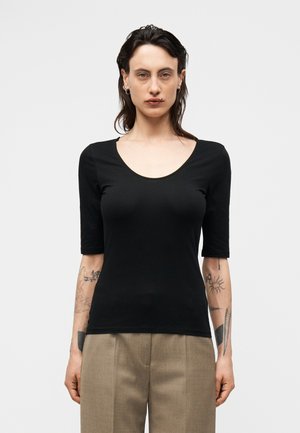 Asmuo su tamsiais, iki pečių siekiančiais plaukais, dėvintis juodą prigludusią palaidinę su apvalia iškirpte ir smėlio spalvos kelnes, rodantis tatuiruotes ant abiejų dilbių.