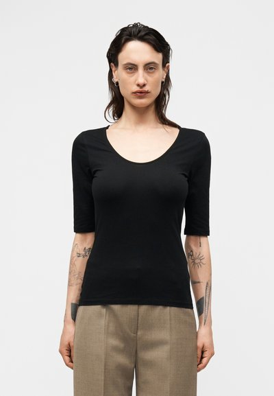 Person with shoulder-length dark hair wearing a fitted black scoop-neck top and beige trousers, showing tattoos on both forearms.