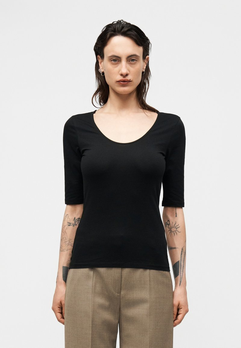 Άτομο με σκούρα μαλλιά μέχρι τους ώμους φορώντας εφαρμοστή μαύρη μπλούζα με στρογγυλή λαιμόκοψη και μπεζ παντελόνια, δείχνοντας τατουάζ και στα δύο κάτω χέρια.