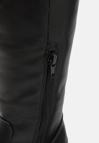 Botte noire en cuir, hauteur genou, avec une fermeture éclair sur le côté et une texture lisse. La fermeture éclair est brillante, ce qui met en valeur le détail de conception global.