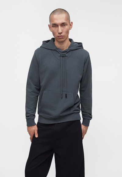 Homme aux cheveux courts portant un sweat à capuche gris foncé avec l'étiquette "ICEBERG" et un pantalon noir, debout devant un fond blanc uni.