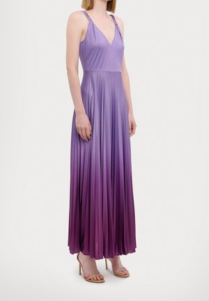 Femme portant une robe maxi plissée dégradée violette sans manches avec de fines bretelles et des sandales à talons hauts beiges, debout devant un fond uni.
