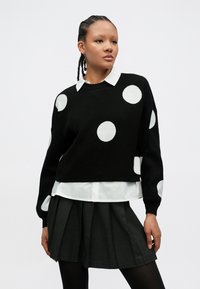 Camisola preta de malha com bolinhas brancas, gola redonda e mangas compridas, utilizada sobre uma camisa branca com colarinho e uma saia plissada cinzenta.