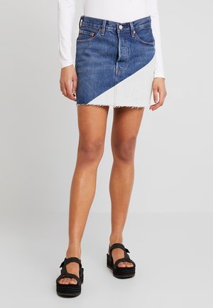 Femme portant une mini-jupe en jean bicolore avec un panneau blanc diagonal, associée à un haut blanc à manches longues et des sandales compensées noires.