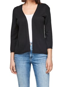 Woman wearing a black open-front cardigan over a white top with light blue jeans, standing against a white background.