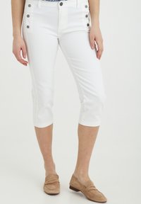Pantaloni capri bianchi dal design aderente, con bottoni decorativi sui lati e una consistenza morbida, abbinati a mocassini in suede beige chiaro.