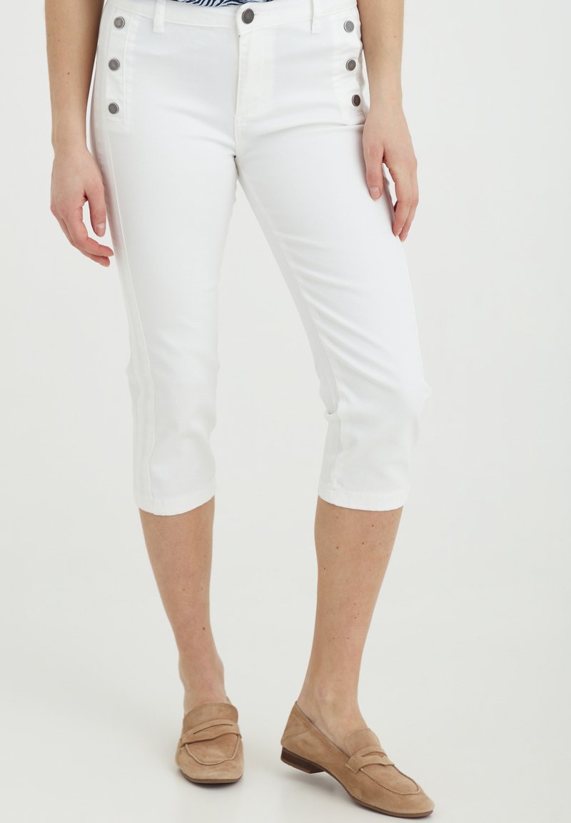 Pantaloni capri bianchi dal design aderente, con bottoni decorativi sui lati e una consistenza morbida, abbinati a mocassini in suede beige chiaro.