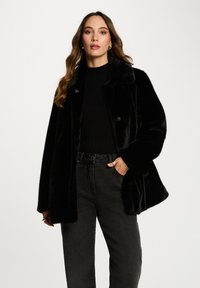 Cappotto in pelliccia sintetica nera con colletto, chiusura a bottoni e vestibilità ampia. Abbinato a jeans neri a vita alta. Texture soffice, design elegante.