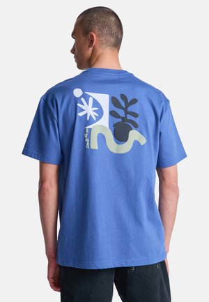 Mann in blauem T-Shirt mit abstrakter Kunst von Pflanze, Stern und wellenförmiger Form auf dem Rücken, steht vor schlichtem weißen Hintergrund.