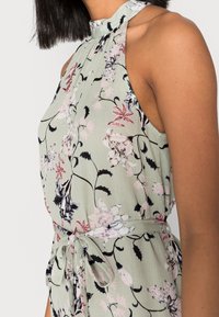 Květinový top s halter krkem vyrobený z lehkého texturovaného materiálu. Má mátově zelené pozadí s růžovými a černými květinovými vzory, stažený v pase.