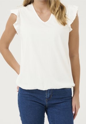 Vrouw in een witte mouwloze top met gerimpelde randen en een spijkerbroek, staand met één hand in de zak tegen een egale achtergrond.