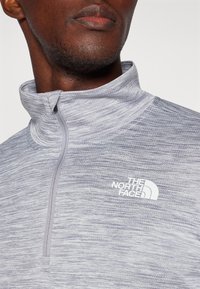 Grå genser med høy krage, halv-zip-design og subtil horisontal tekstur. Har hvit logo på brystet. Mykt stoff.