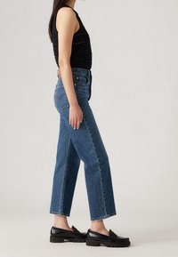 Top negro sin mangas con detalles de corte horizontal, jeans rectos de tiro alto azul y mocasines negros con suelas gruesas.