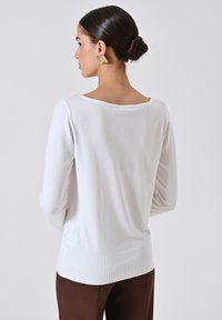 Top bianco a maniche lunghe realizzato in un tessuto morbido e texturizzato. Presenta una scollatura a barco e orlo a coste, abbinato a pantaloni marroni.