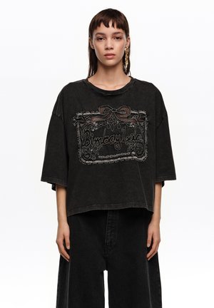 Joven con cabello castaño liso que lleva una camiseta negra desgastada oversized con un diseño floral bordado y texto, combinada con pantalones negros sueltos.