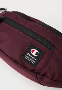Banane en tissu bordeaux avec une fermeture éclair noire, présentant un patch de marque en blanc et rouge. Surface texturée avec un design compact.