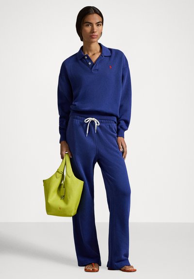 Kraljevsko modra majica z ovratnikom in gumbi, v kombinaciji z ujemajočimi trenirkami z vrvico za zavezovanje. Poudarek z živordečo zeleno torbo za na ramo.